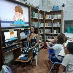 Professora conduz aula de inglês com apoio de vídeo educativo na biblioteca de Copacabana, Rio de Janeiro.